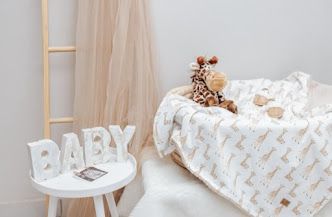 Cozy nursery corner with a giraffe blanket, stuffed toy, wooden ladder, sheer canopy, and BABY letter sign on a white table.