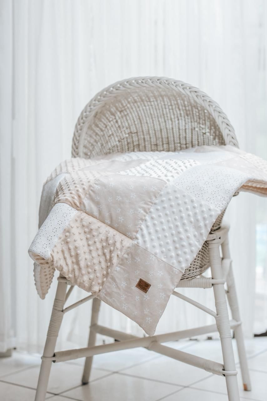 A white minky textured blanket is draped over a light-colored wicker chair against a white curtained background.