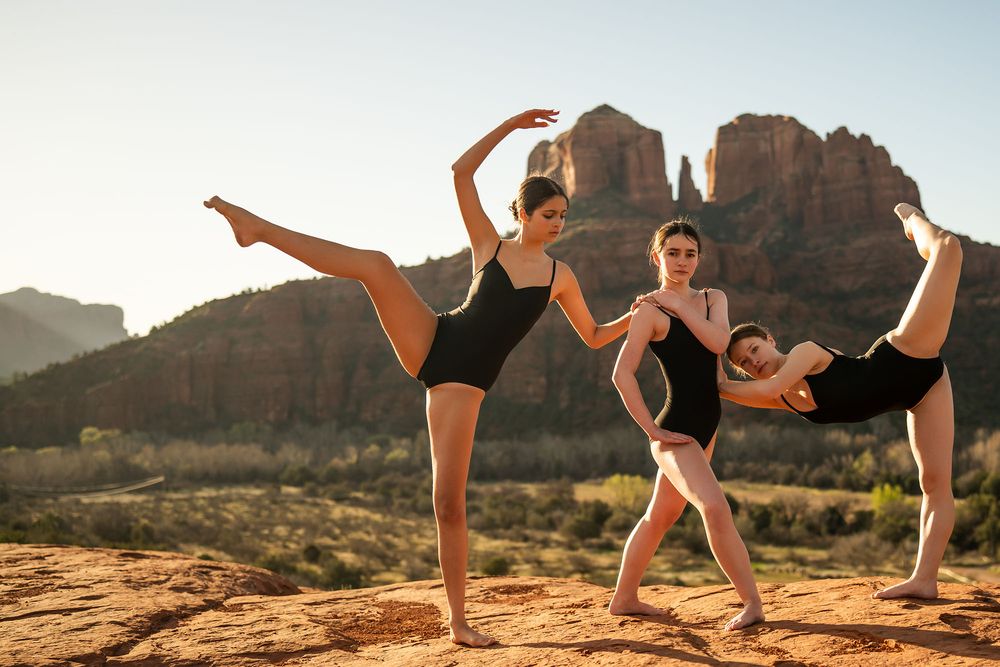 Three ballet dancers performing graceful high kicks outdoors in Sedona, Arizona with red rock formations in background.