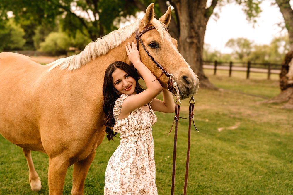 Happy girl in white lace dress smiling while embracing a palomino horse in a lush green pasture.