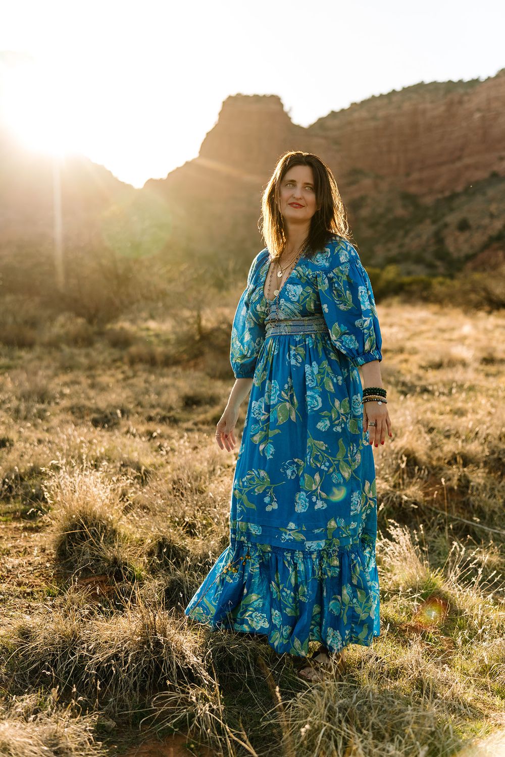 Woman in blue floral maxi dress standing in desert field with red rock buttes at golden sunset.