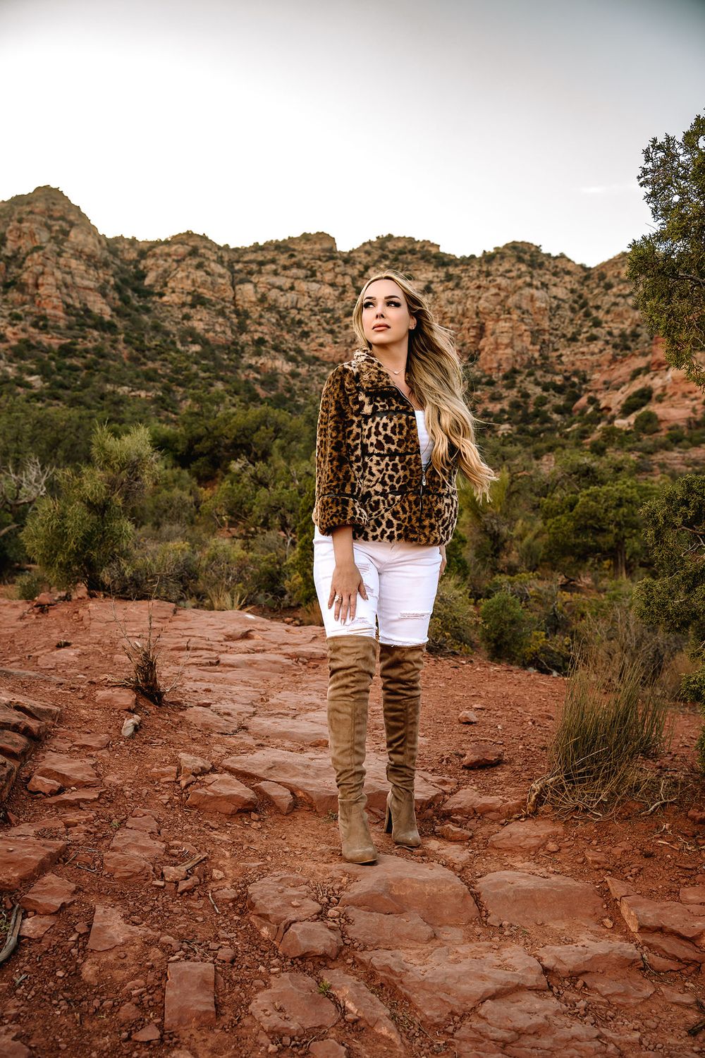 Woman in leopard jacket, white jeans, and tan boots stands on red rock trail with desert mountains backdrop.