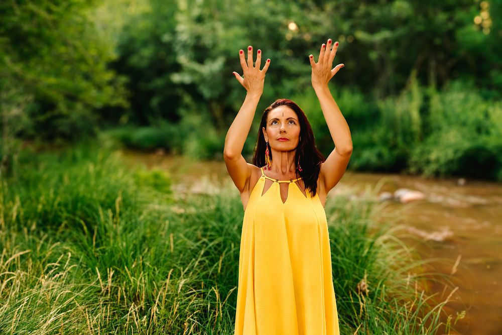 Woman in yellow dress raises hands outdoors near lush green river bank in serene nature setting.