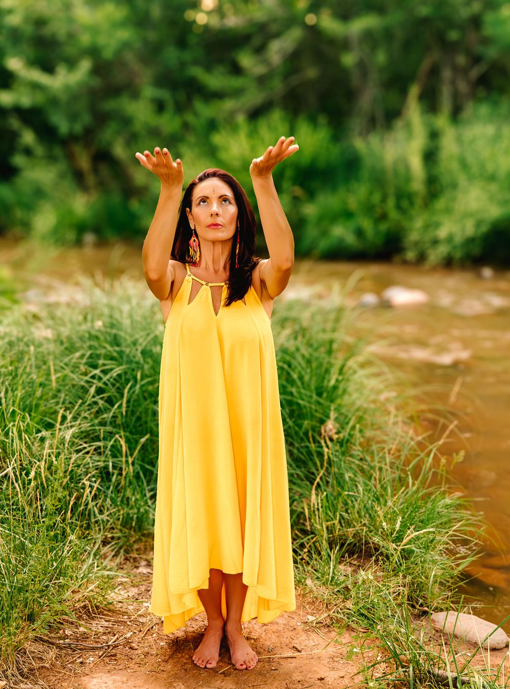 Woman in yellow dress raises hands skyward barefoot by a creek surrounded by lush green nature.