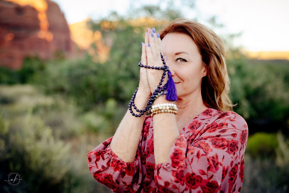 Woman in floral dress holding mala beads in prayer pose with Sedona red rocks at sunset.