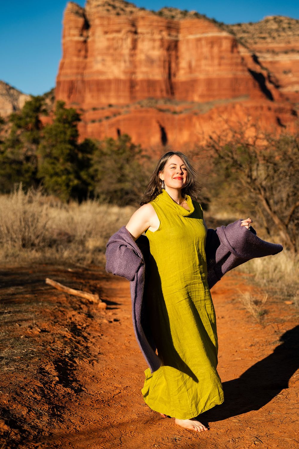 Woman in yellow dress with purple jacket posing before red rock formations in Sedona.