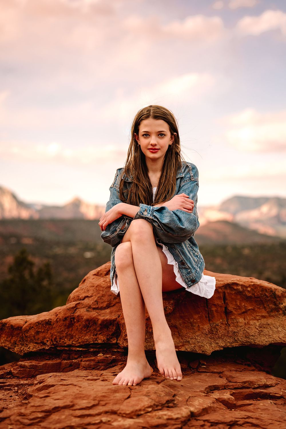 Young woman in denim jacket sitting barefoot on red rocks with mountain views at sunset.