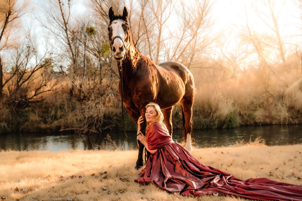 Elegant portrait in flowing red dress with majestic horse against sunset backdrop.