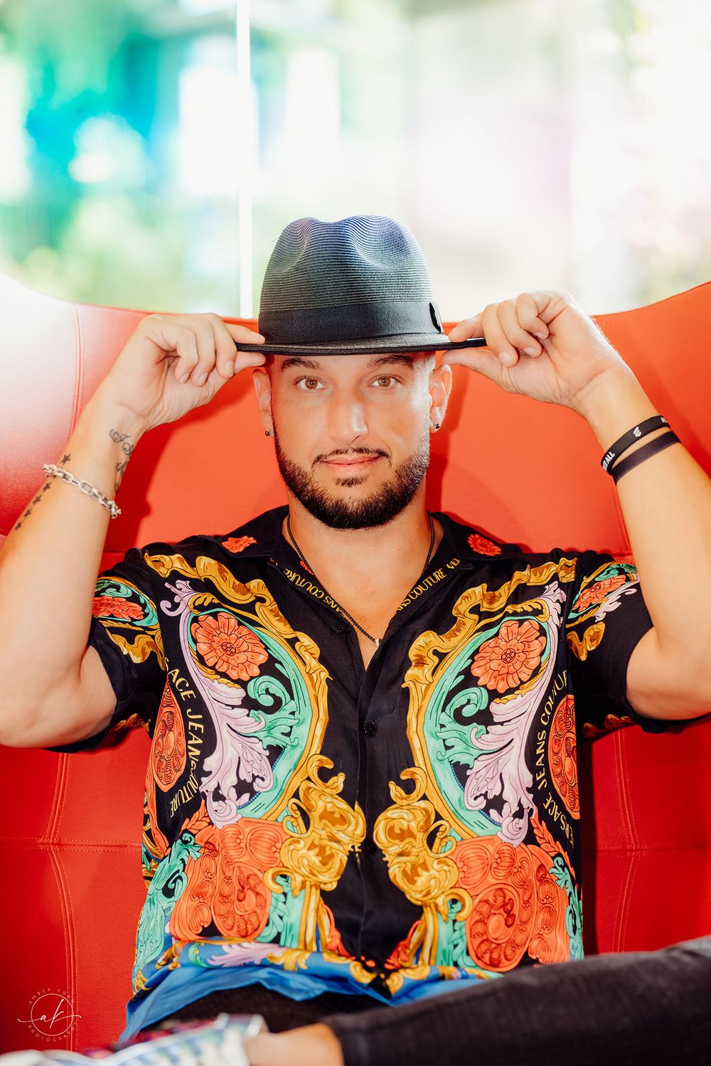 Man in colorful baroque shirt and gray fedora hat posing in red chair with confident expression.