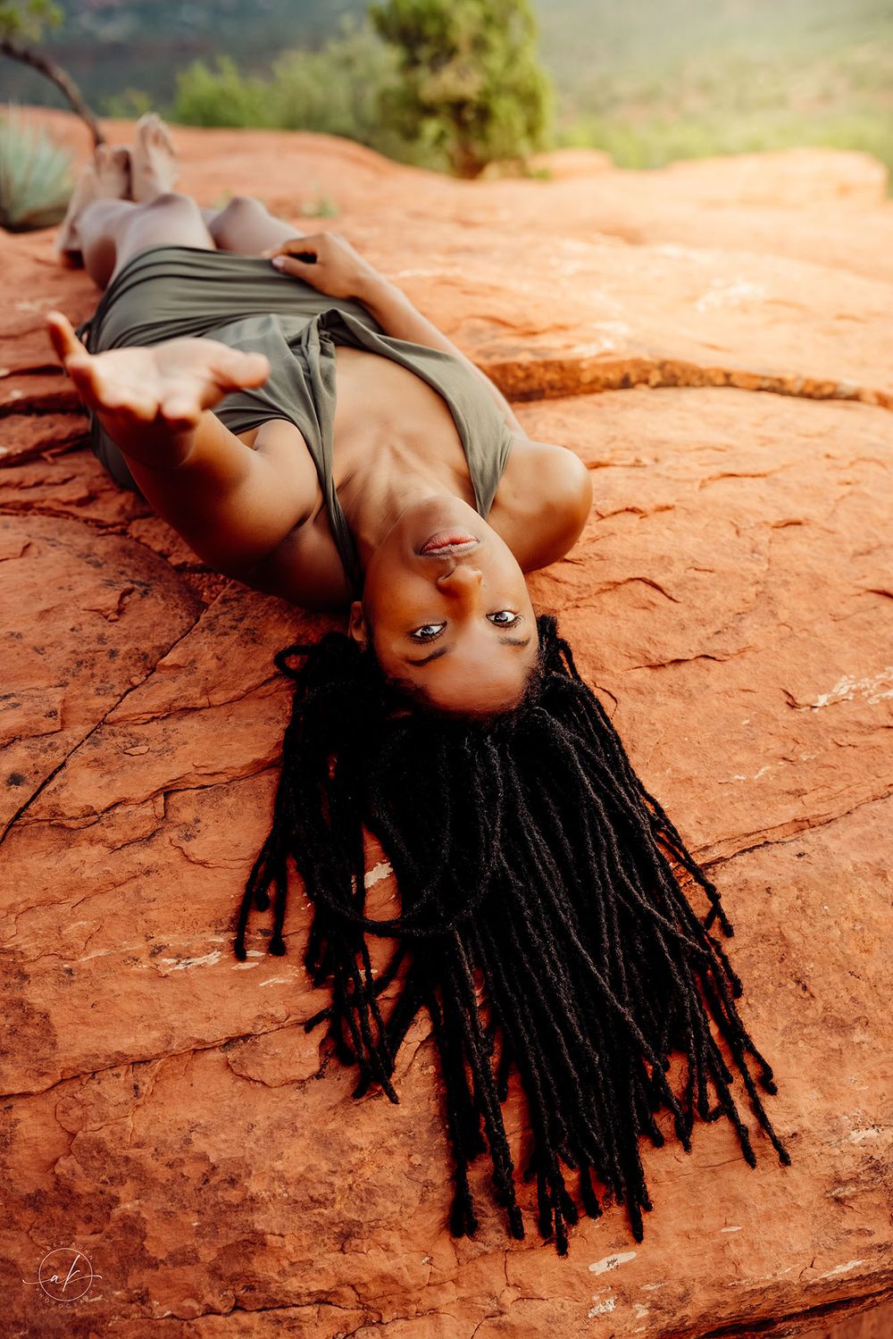 Woman with long braids lying on red rock formation in a sage green dress, viewed from above in natural desert setting.