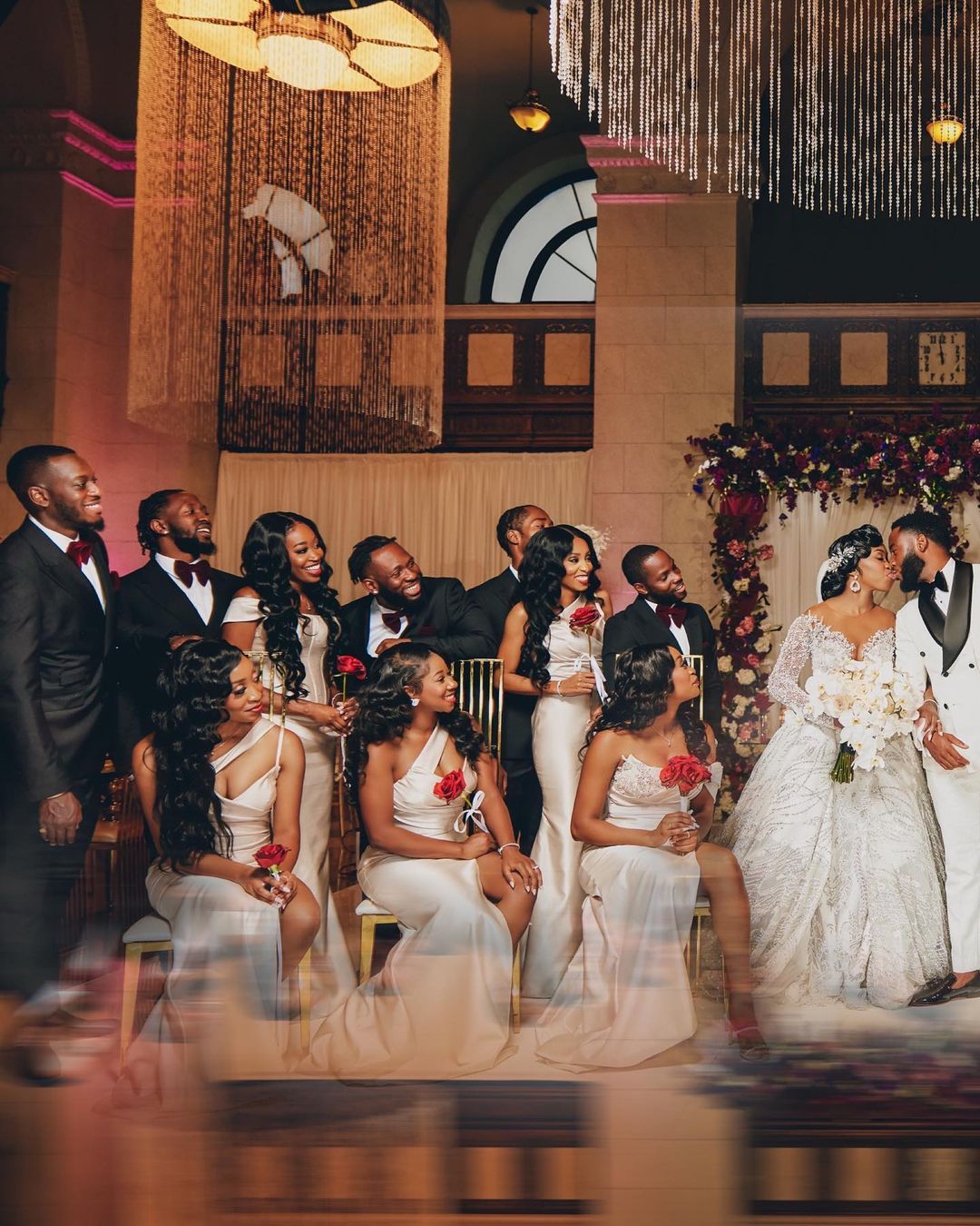 Wedding party in formal attire gathered in elegant venue with chandelier lighting.
