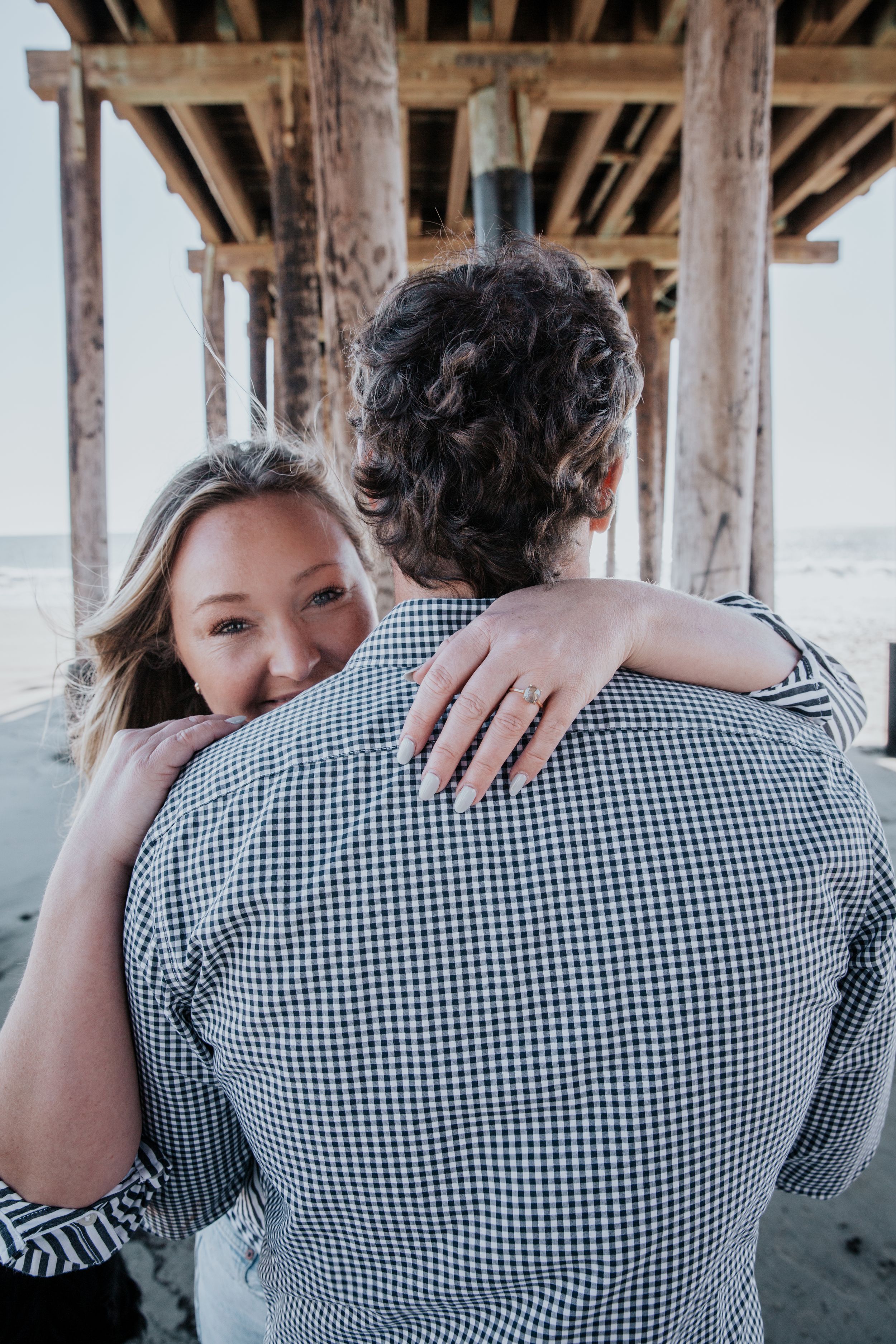 A couple embraces under a pier, with a close-up view showing a gingham button-down shirt and an engagement ring.