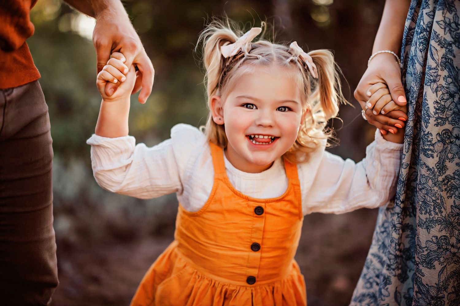 A joyful child in an orange jumper dress holds hands with adults while playing outdoors.