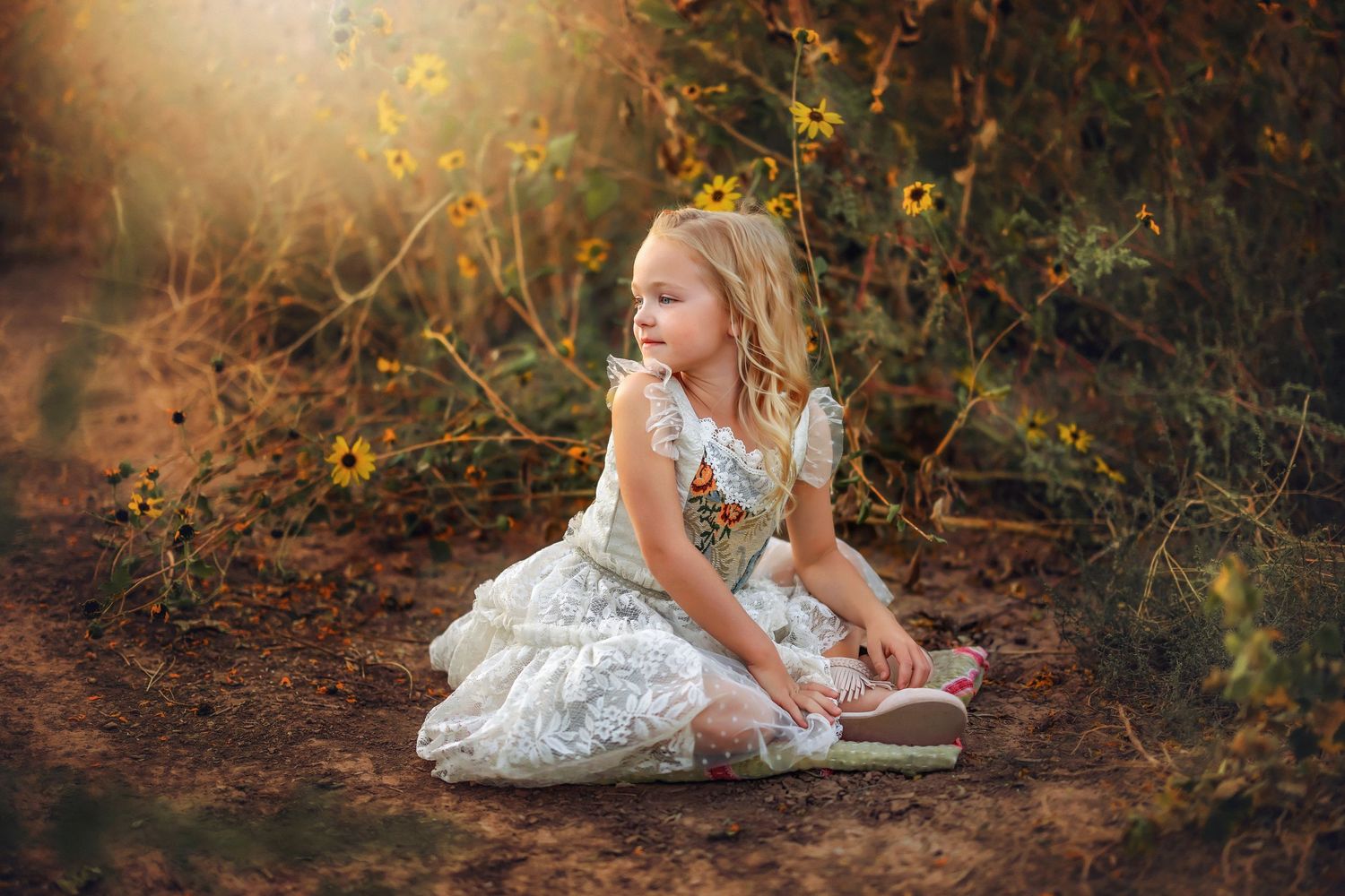 A child in a white dress sits in dappled sunlight among yellow wildflowers.