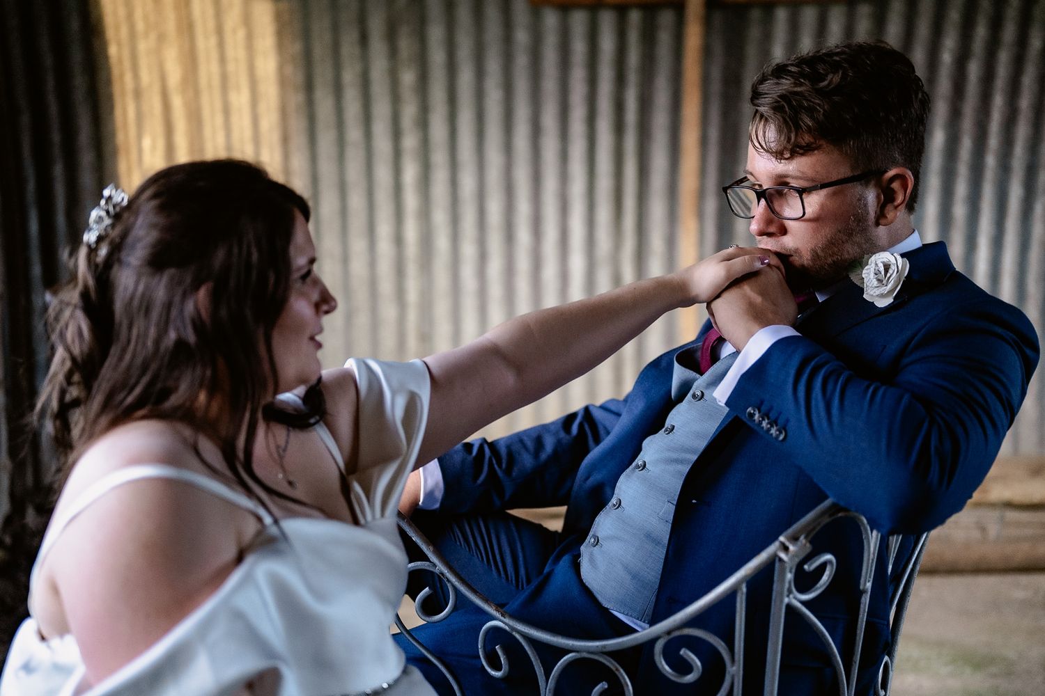 A bride and groom share an intimate moment while sitting on a vintage metal chair against a rustic corrugated wall.