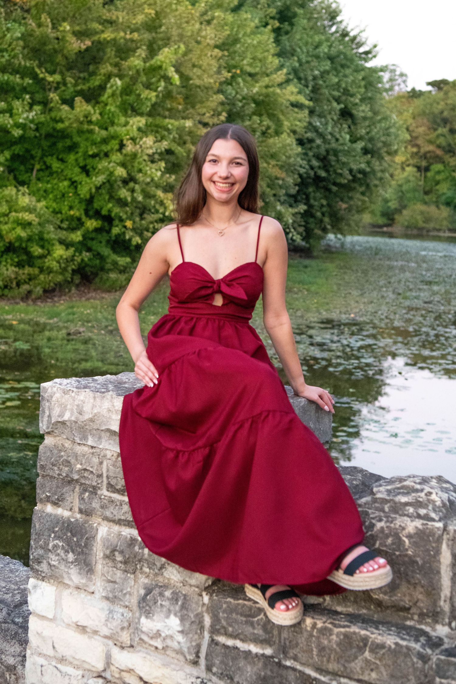 Someone in a flowing burgundy dress sits on a stone wall by a peaceful pond surrounded by lush green trees.