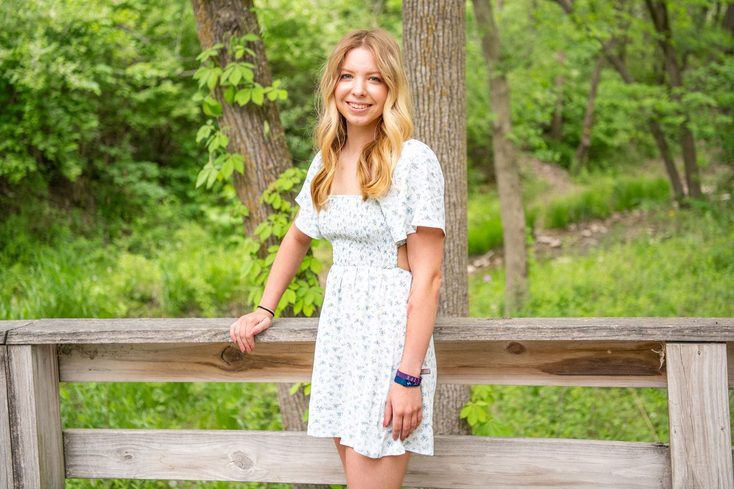 Person in white floral dress poses along wooden fence in green forest setting.