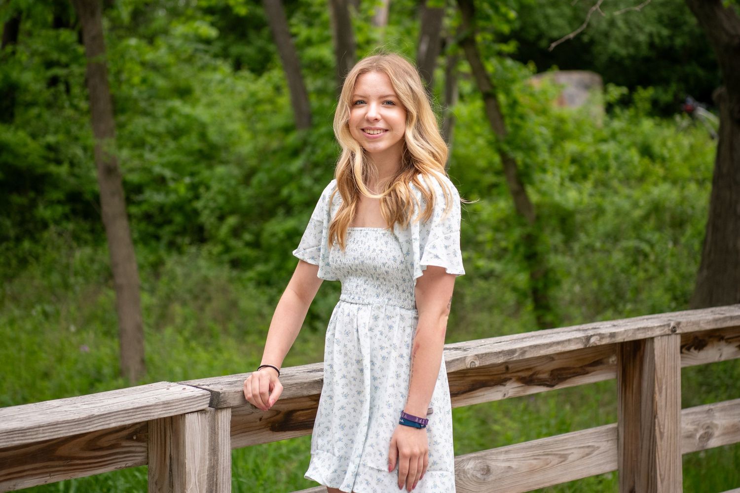 A white floral dress is showcased in multiple angles against a wooden fence and green background.