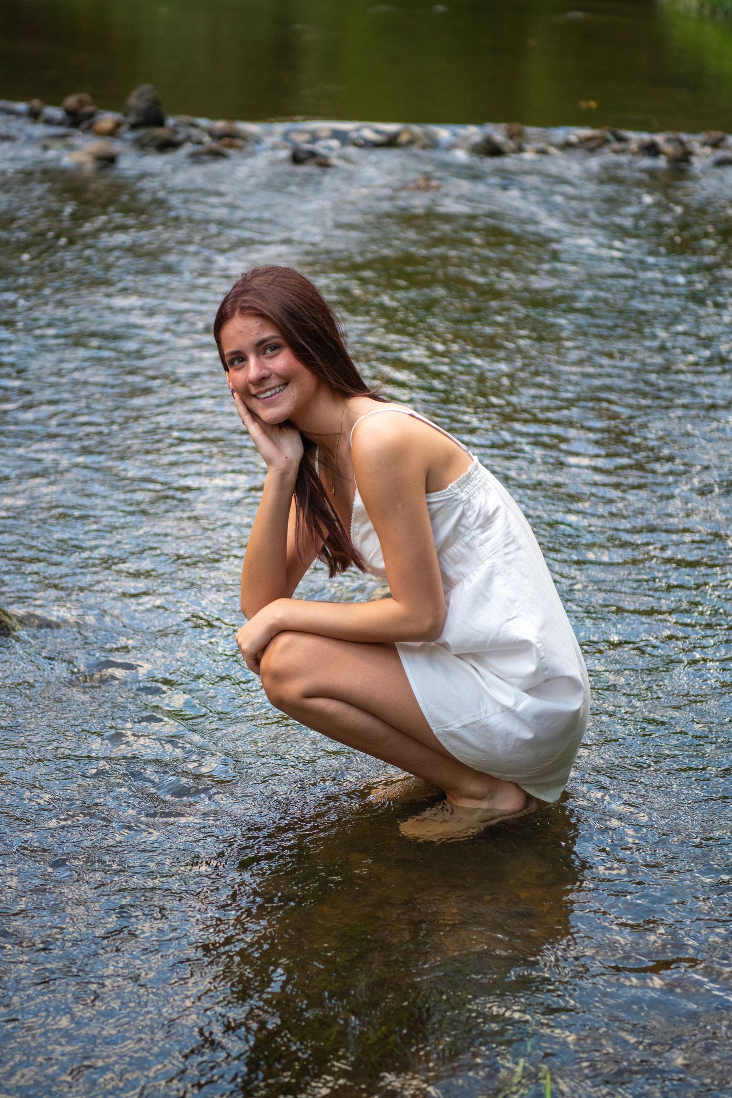 Someone in a white sundress crouches by a rocky stream on a summer day.