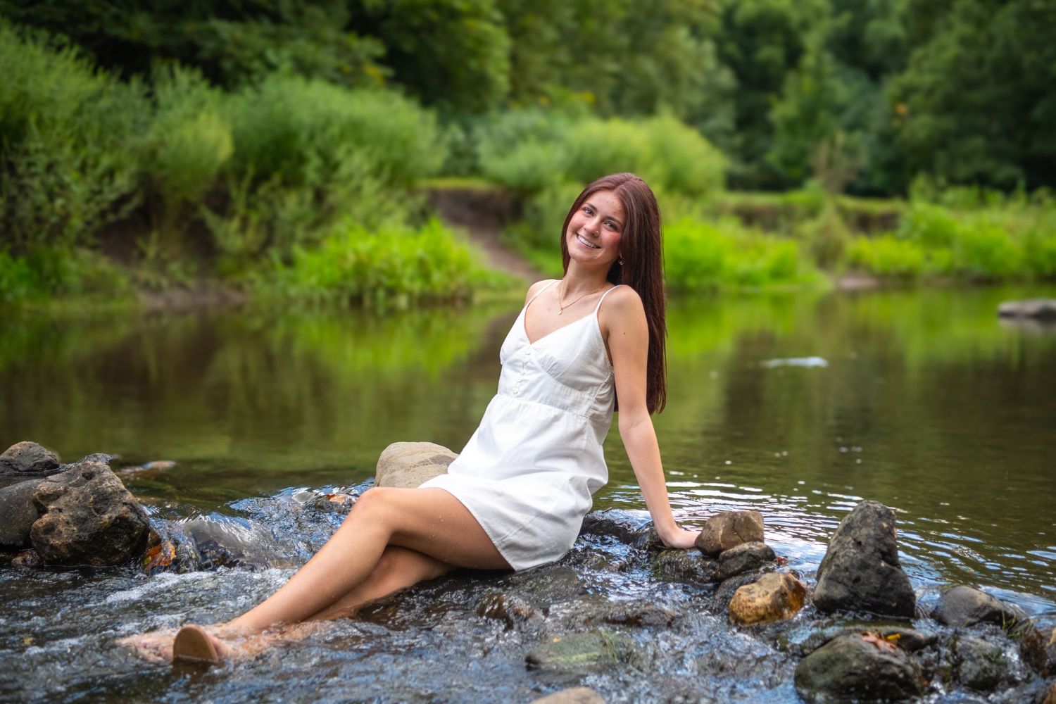 Woman in white dress posing by a peaceful stream on mossy rocks in a serene forest setting.