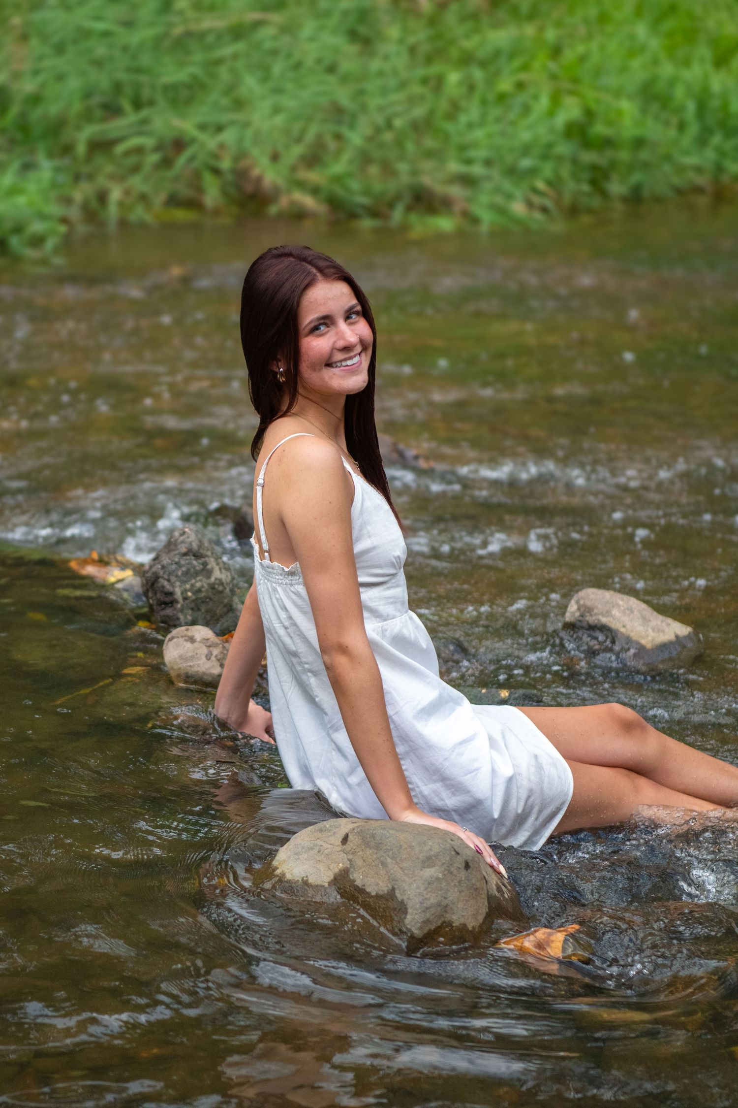Person in white summer dress sits on rocks in flowing stream with green foliage in background.