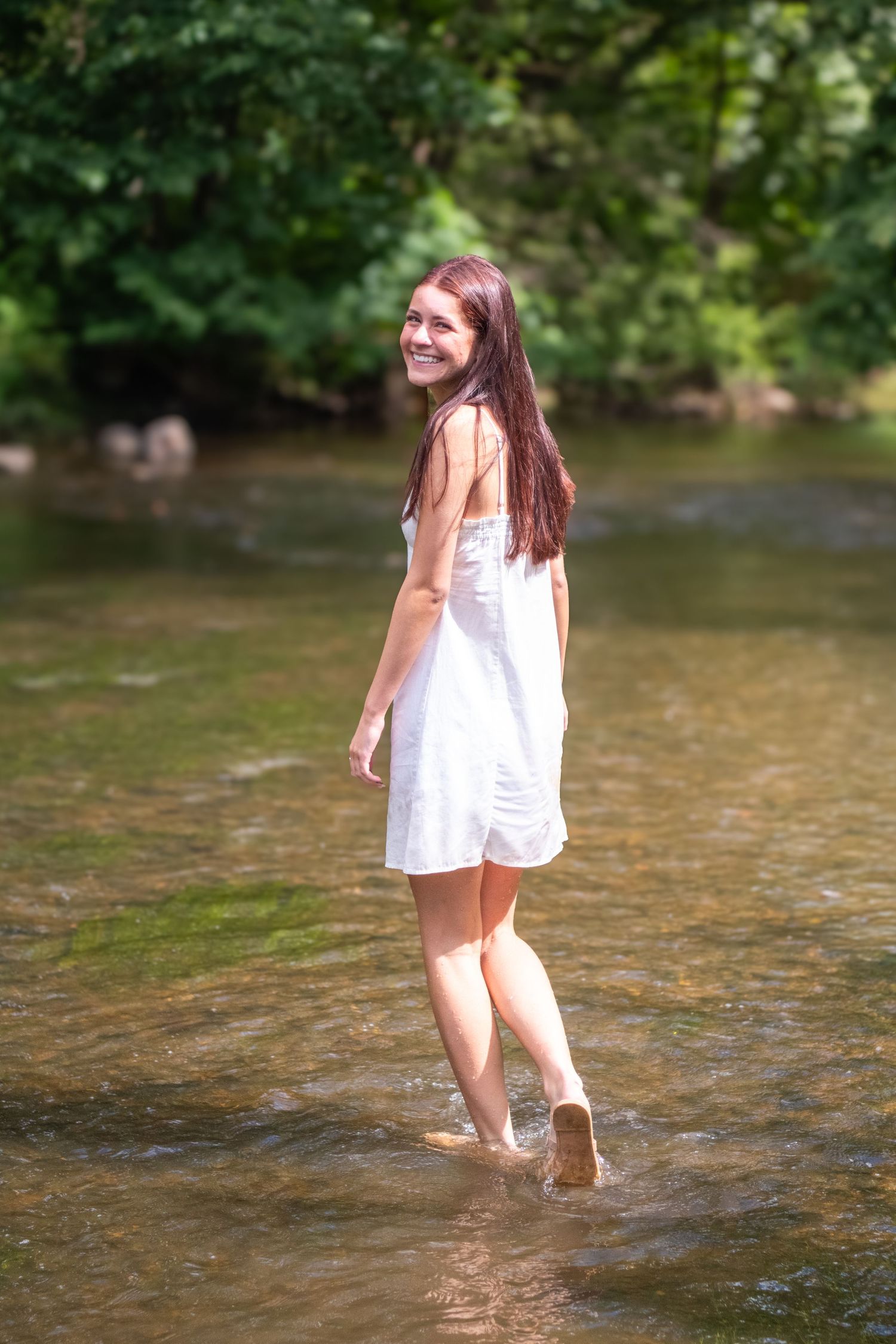 Person in a white sundress walking through shallow creek water on a sunny summer day.