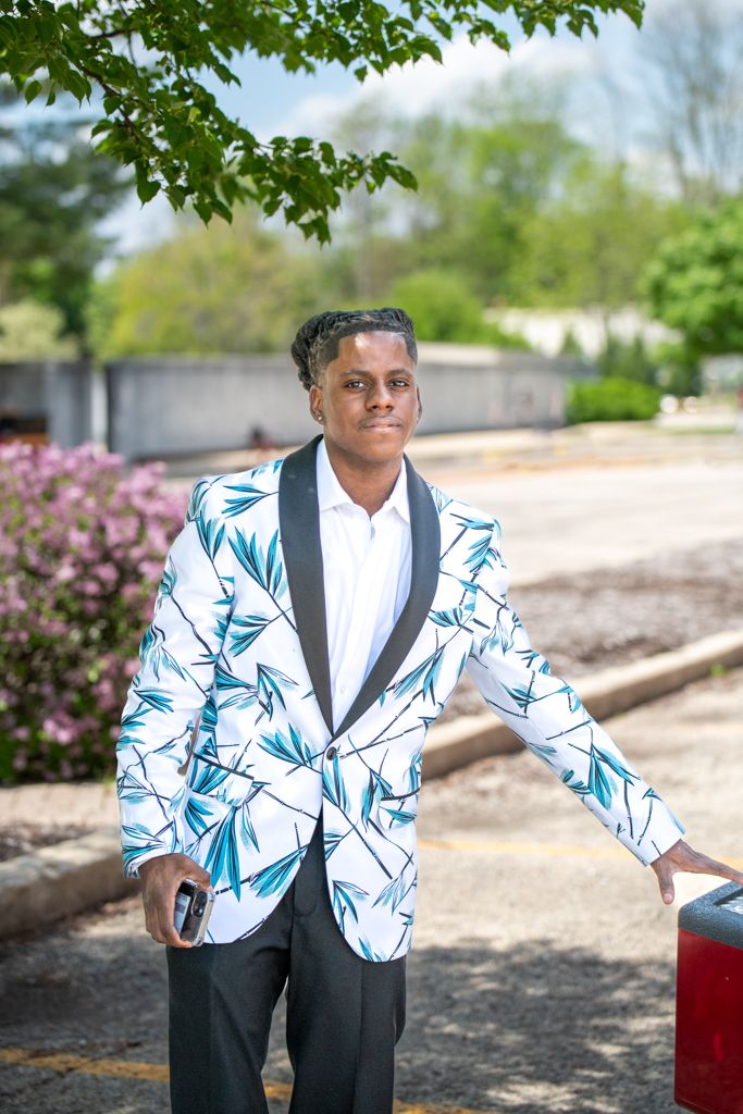 A person in a stylish white and blue patterned blazer poses against a natural outdoor backdrop.