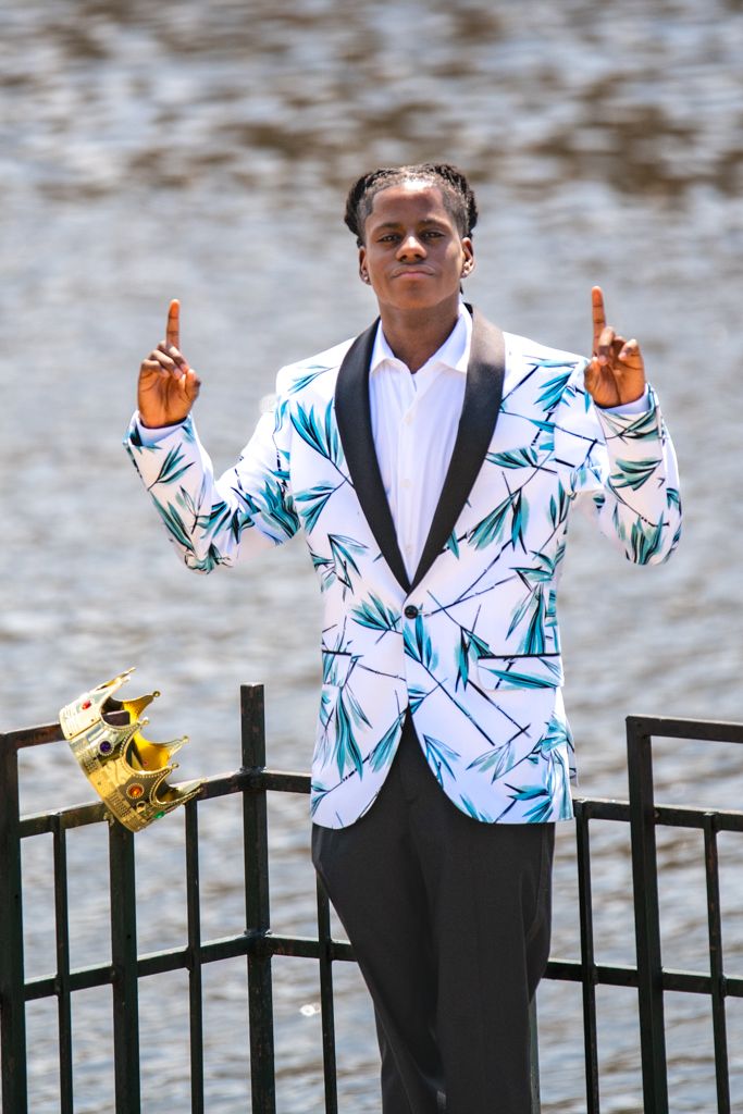 Someone in a striking white blazer with blue designs poses by a railing near water.