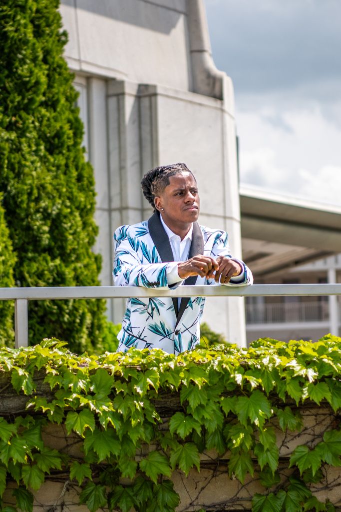 A person wearing a patterned white and blue blazer leans on a wall covered in green ivy.
