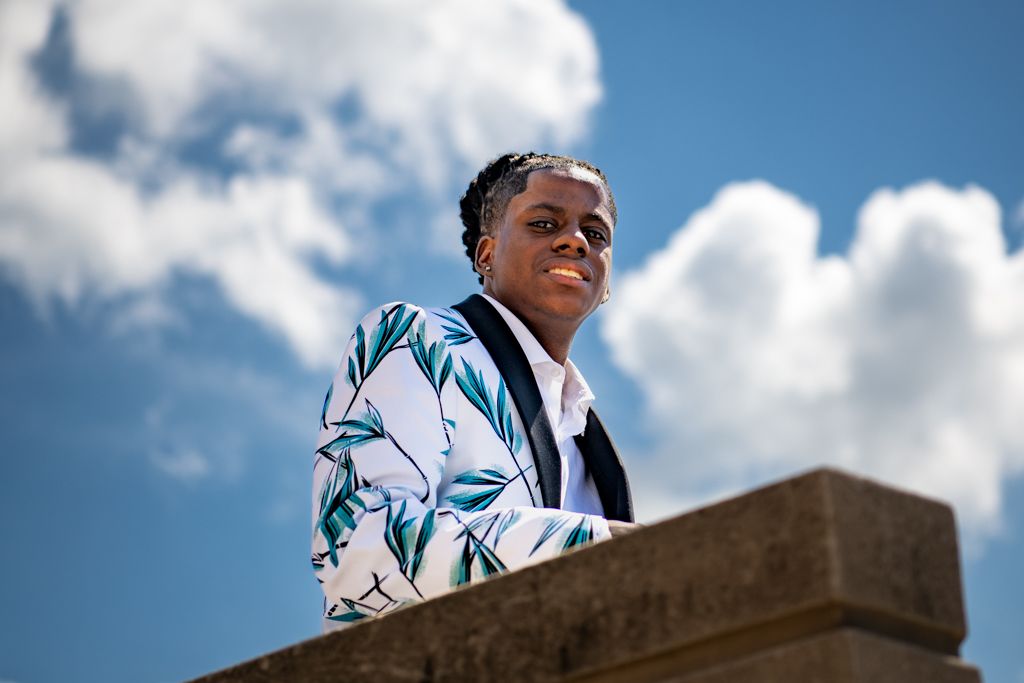 A figure in a white patterned blazer is photographed against a cloudy blue sky.