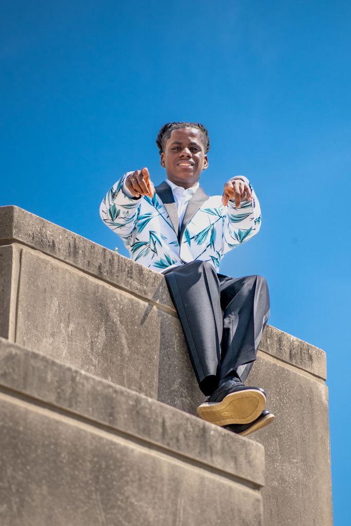 Someone in formal attire with a blue and white jacket sits atop a concrete wall against blue sky.