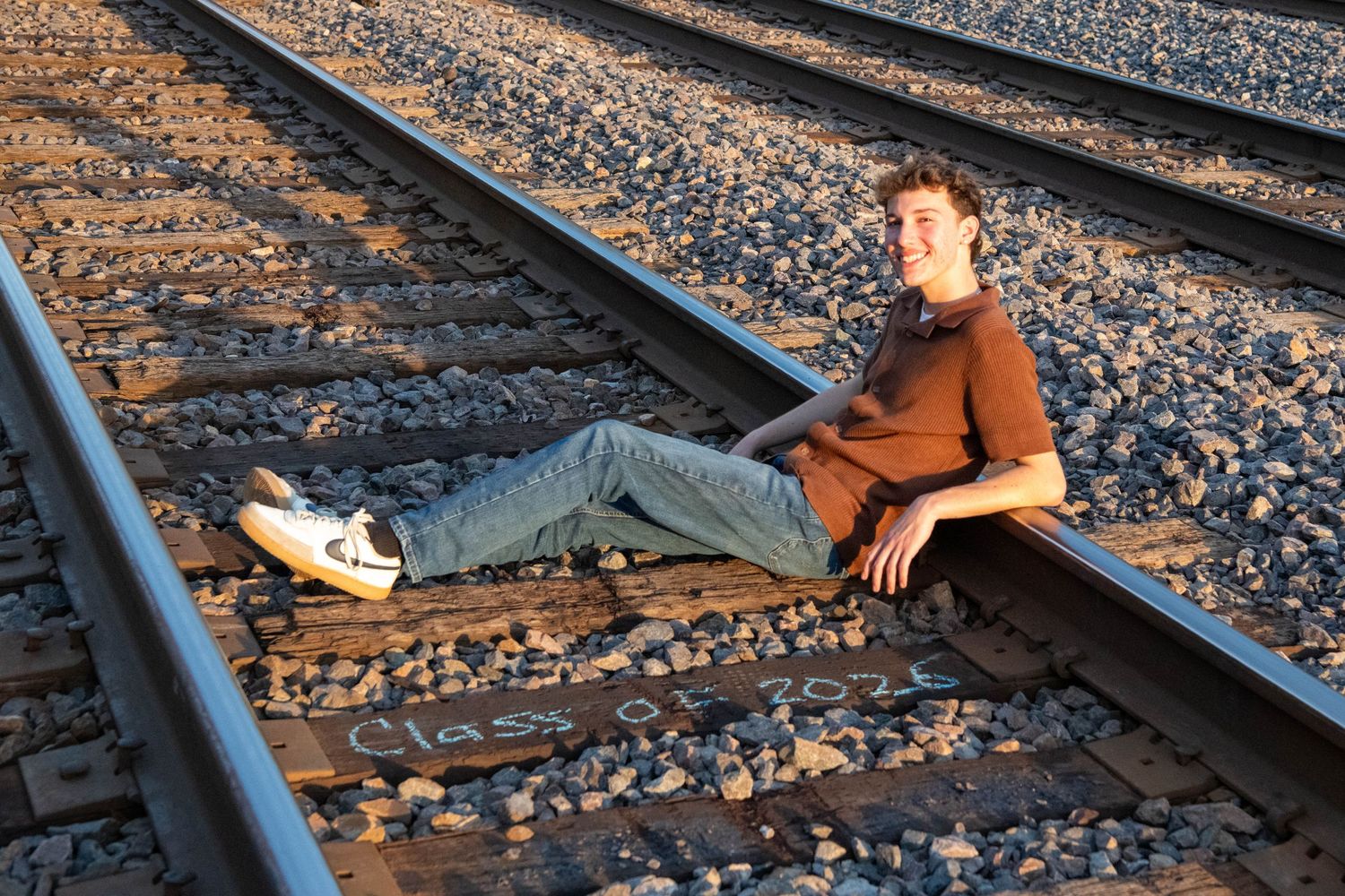 Person in brown jacket sits on railroad tracks during golden hour lighting creating a moody atmospheric scene.