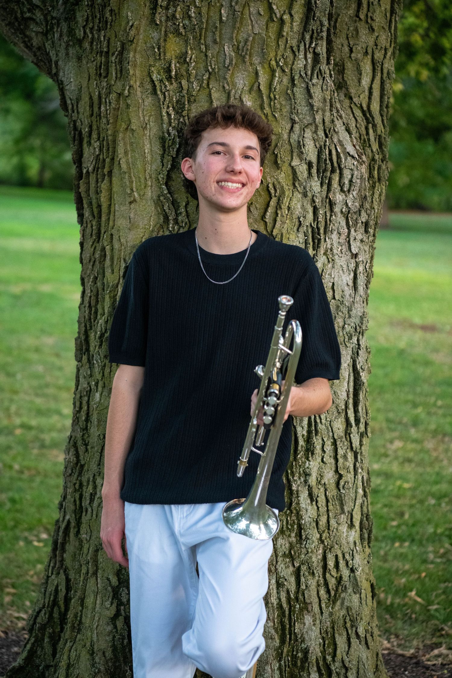 A student poses with their trumpet against a large tree trunk in a park setting wearing black and white attire.