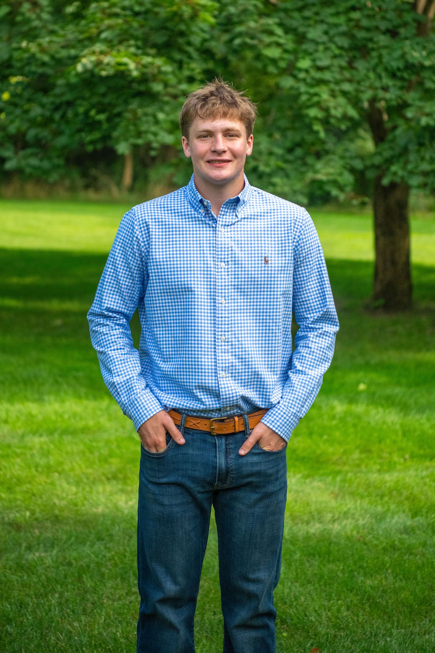 Portrait of someone in blue checkered shirt and jeans standing on grass with trees in background.