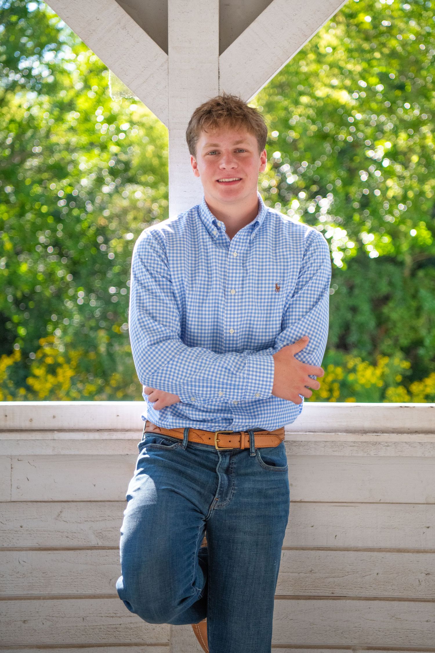 Person in blue gingham shirt and jeans sits casually on white bench with green foliage in background.