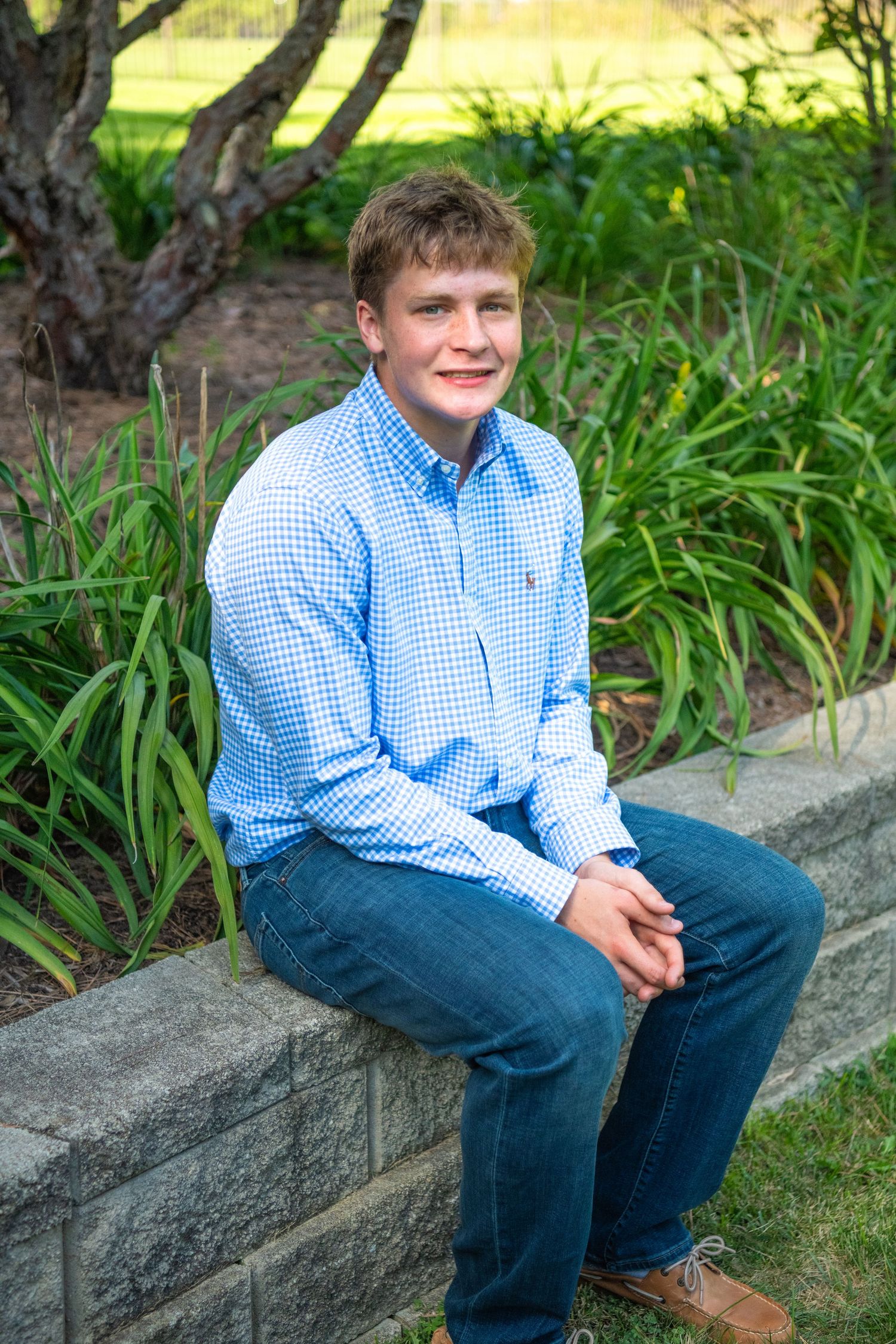 A person wearing a blue gingham shirt and jeans sits casually on a stone wall surrounded by green foliage.