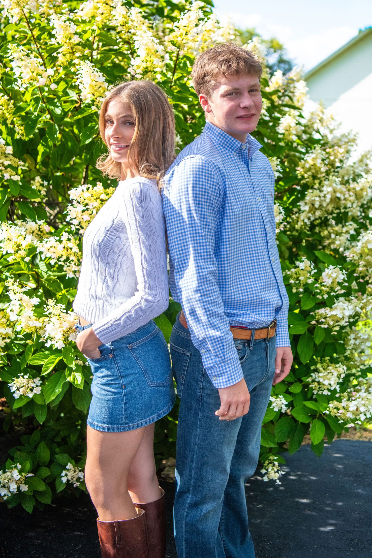 Two people in casual button-down shirts and jeans pose back-to-back against a flowering bush on a sunny day.