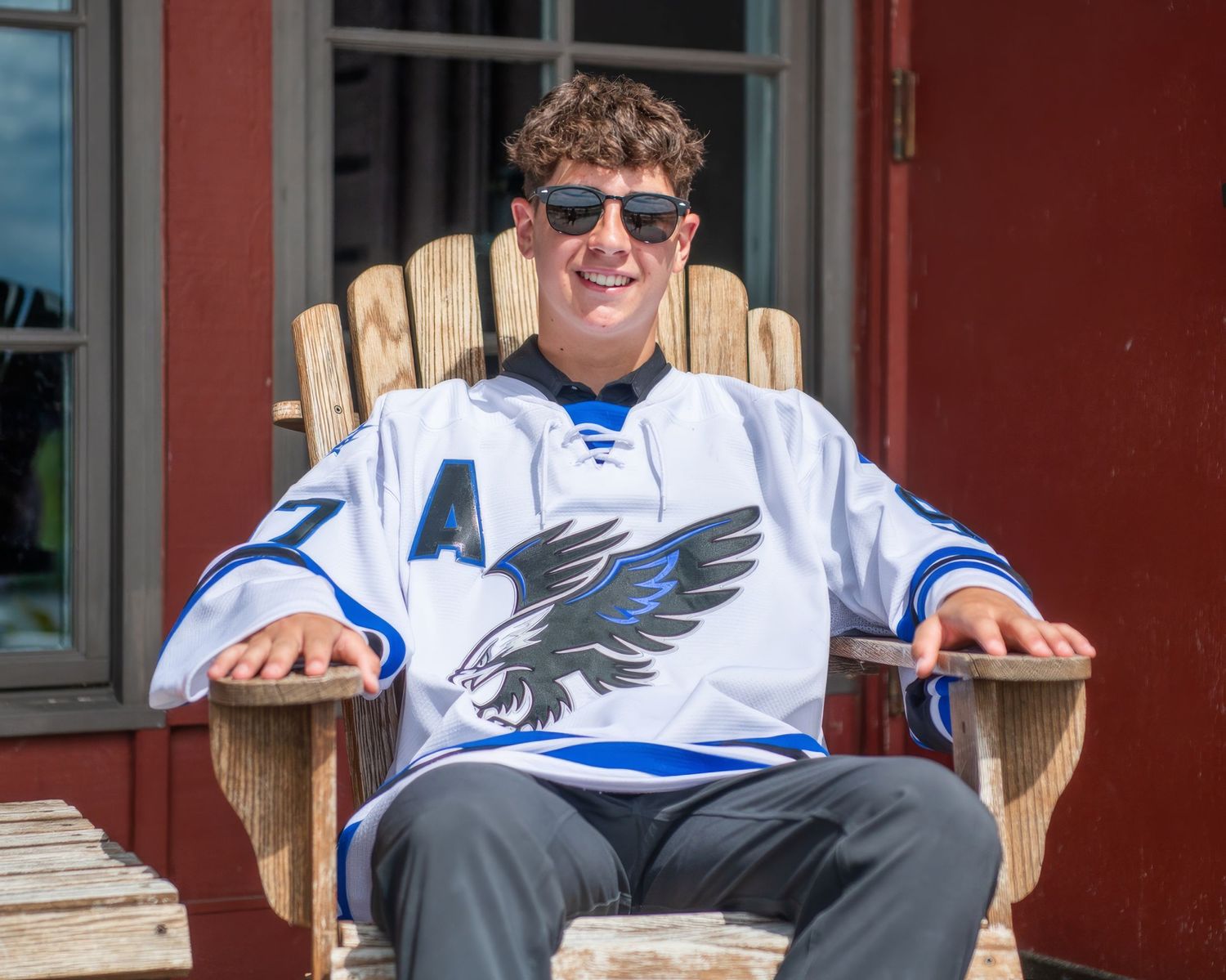 Student in sports jersey seated on wooden bench against red building exterior.