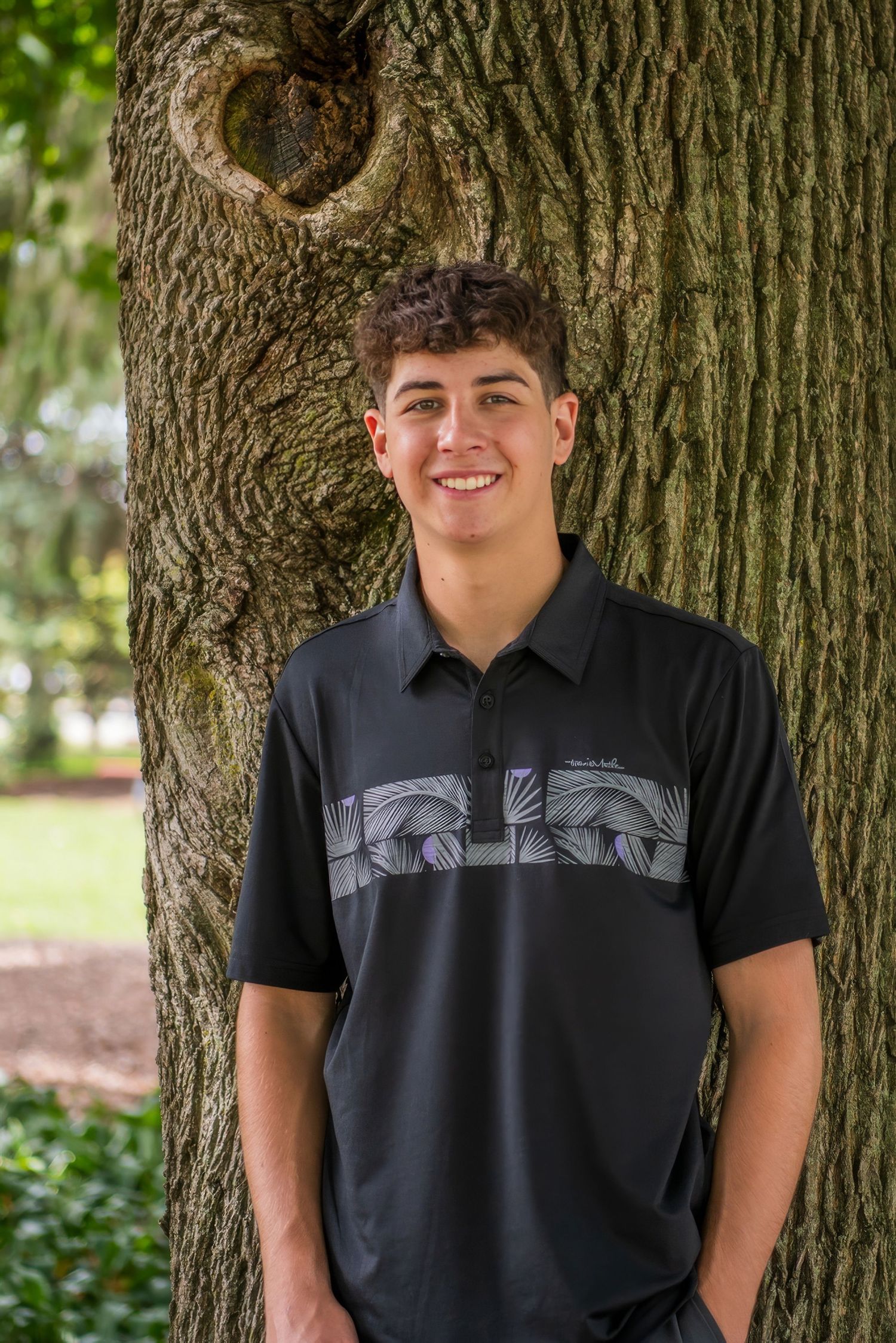 Person in black t-shirt posing against large tree trunk in park setting.