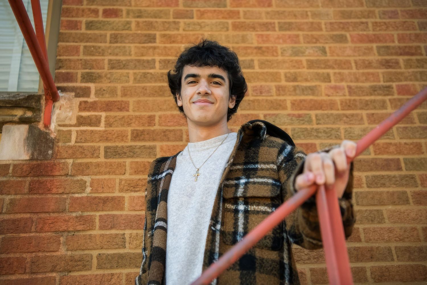 Young person in plaid flannel and white t-shirt leans against brick wall with red railing.