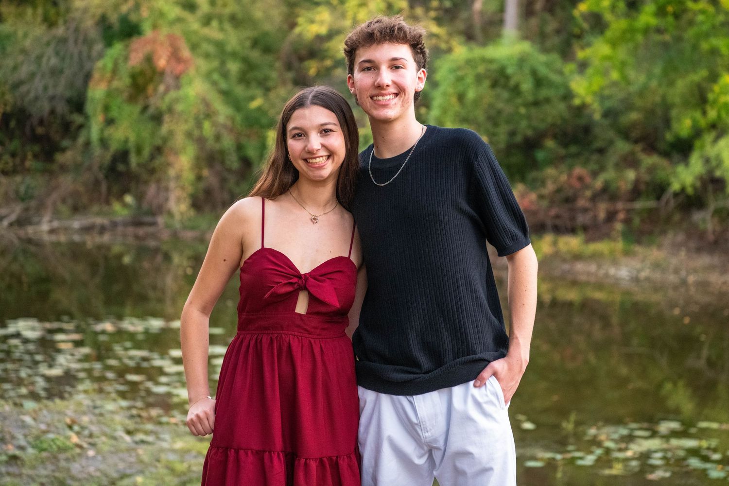 A couple in burgundy and black attire poses together by a scenic pond surrounded by lush greenery.
