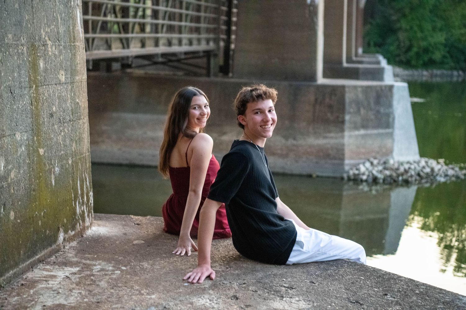 Two people sit together on a stone ledge by the water on a summer day near a bridge.