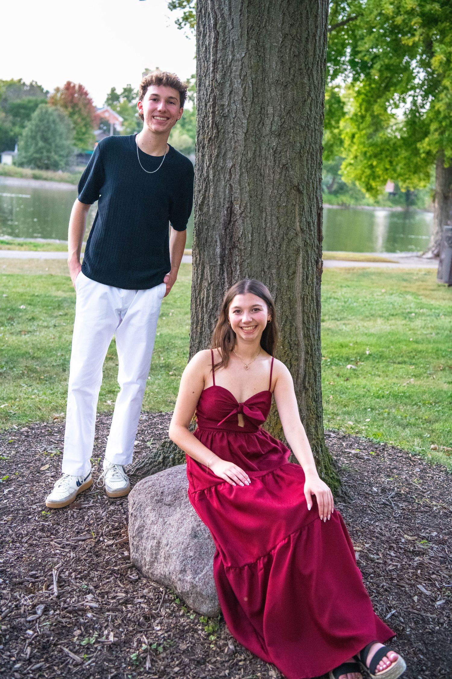 Two people pose by a large tree trunk near a lake, with one standing and one sitting on a rock in a burgundy dress.