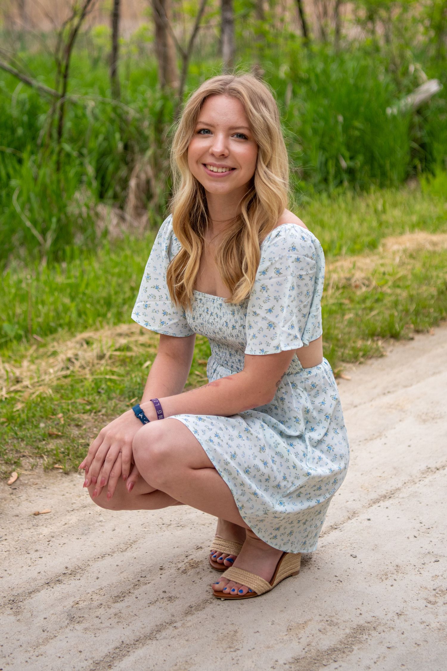 Person in light patterned dress crouches on dirt path surrounded by spring greenery.