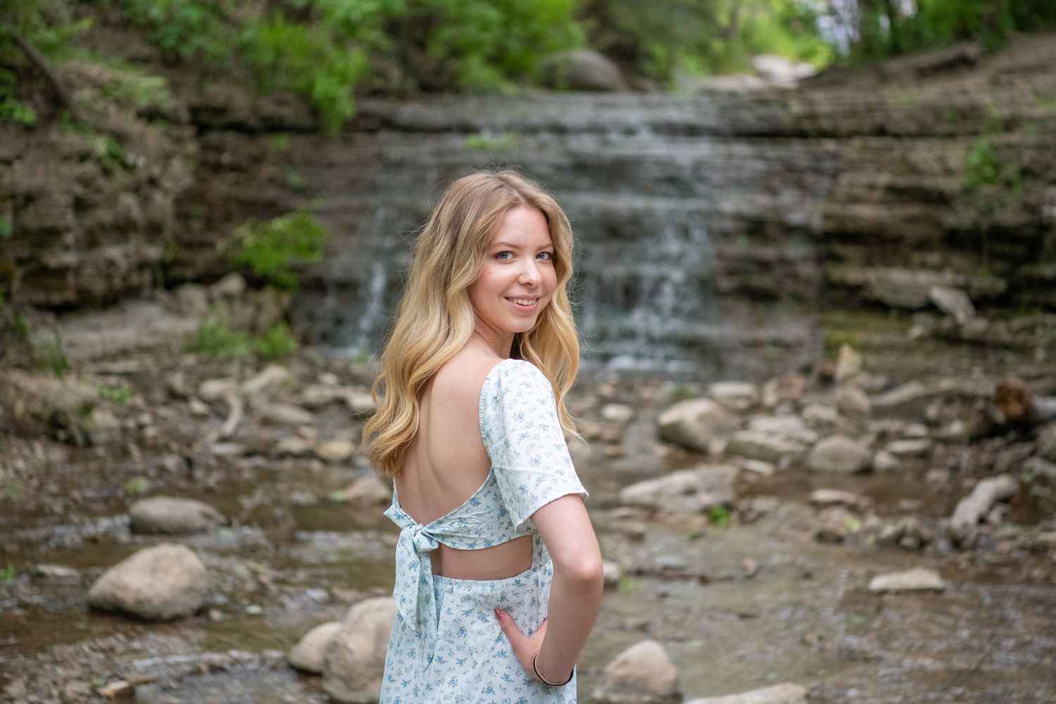 Person in blue dress poses near rocky waterfall with natural stone formations.