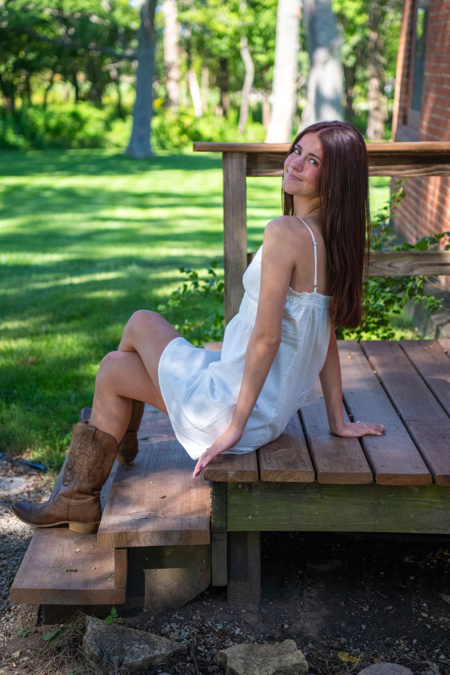 A person in a white dress reclines on rustic wooden deck steps with brown cowboy boots in a sunny garden setting.