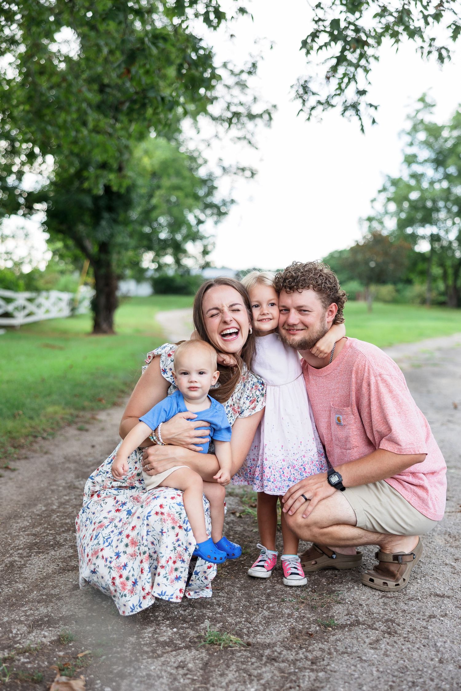 A joyful family of four poses together on a dirt path surrounded by green trees on a sunny day.