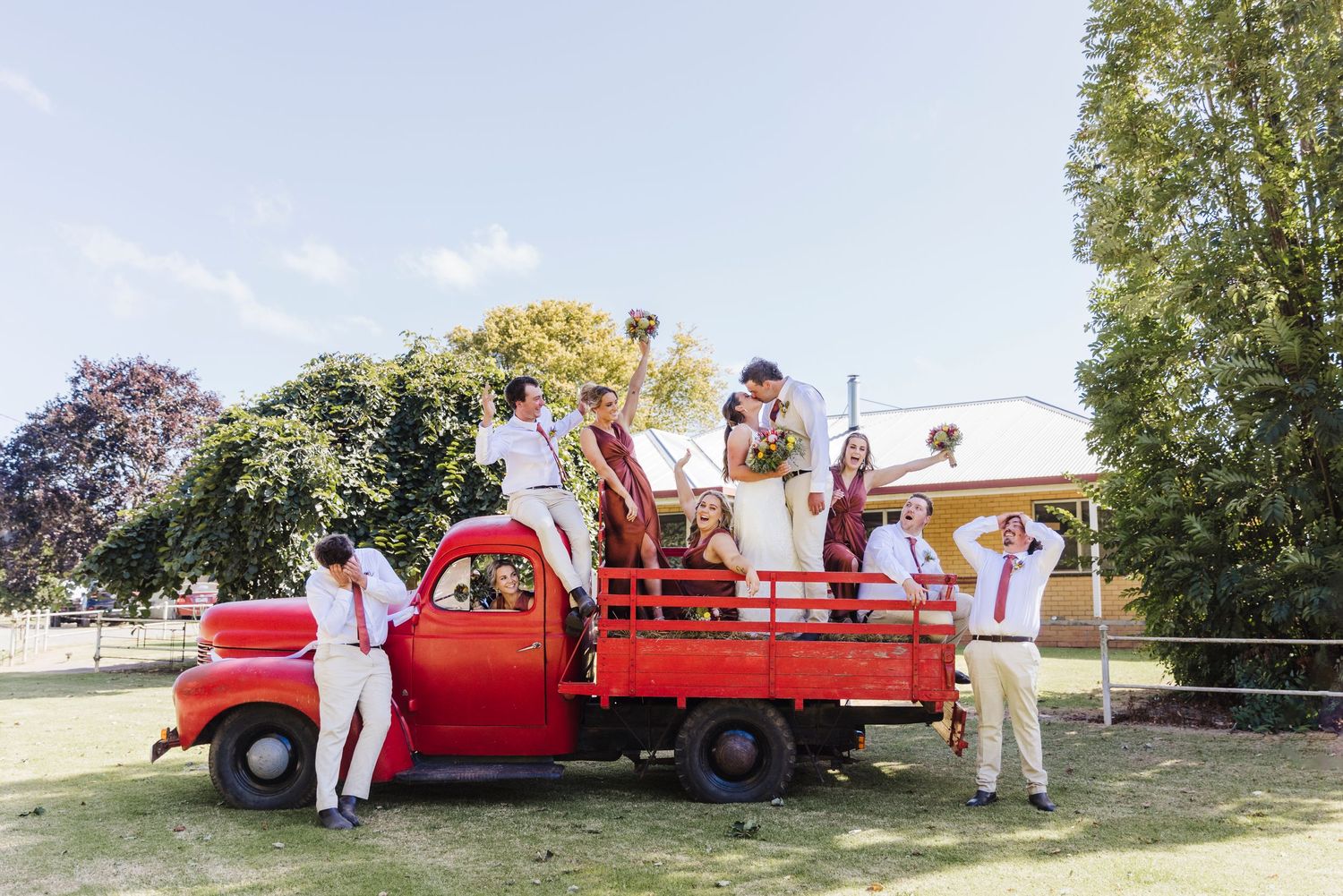 Wedding party posing on a vintage red pickup truck in a rustic outdoor setting.