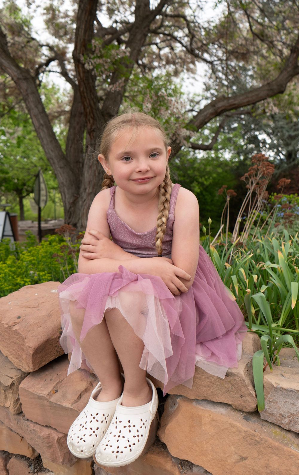 Young girl in pink tulle dress with braids sits on stone wall in lush garden setting.