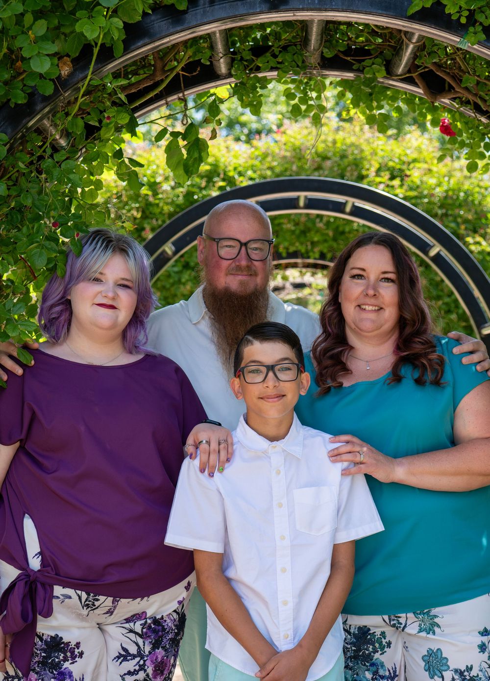 Family of four smiling under a garden archway surrounded by lush greenery on a sunny day.